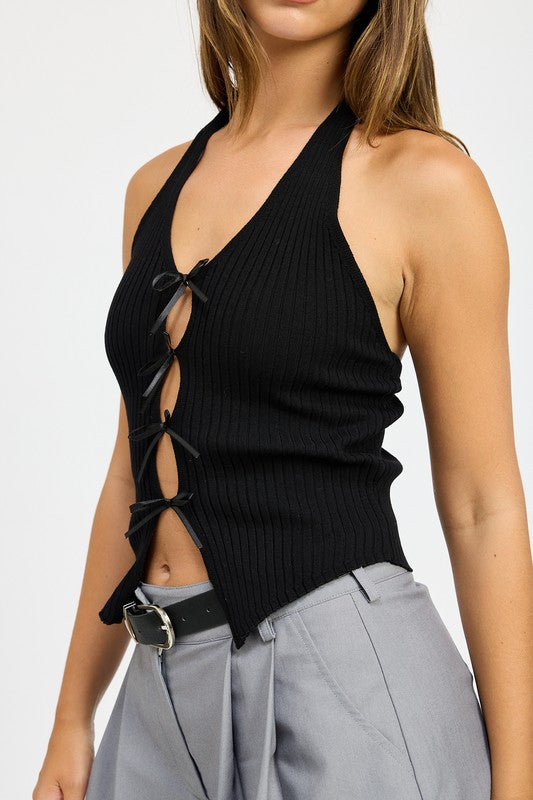 A woman wearing the HALTER NECK TOP WITH BOW DETAILS, a red halter top with black ribbon ties made from 70% viscose and 30% nylon, along with a black headband stands in front of a plain background.