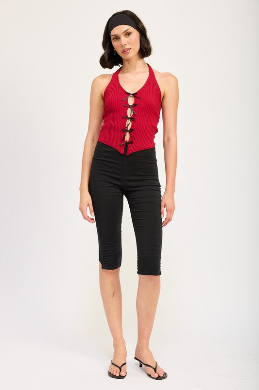 A woman wearing the HALTER NECK TOP WITH BOW DETAILS, a red halter top with black ribbon ties made from 70% viscose and 30% nylon, along with a black headband stands in front of a plain background.