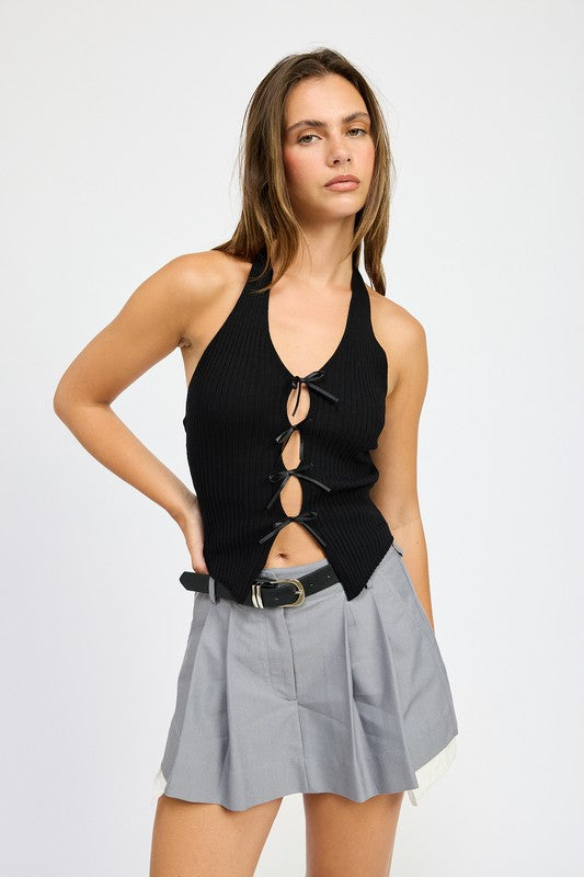 A woman wearing the HALTER NECK TOP WITH BOW DETAILS, a red halter top with black ribbon ties made from 70% viscose and 30% nylon, along with a black headband stands in front of a plain background.