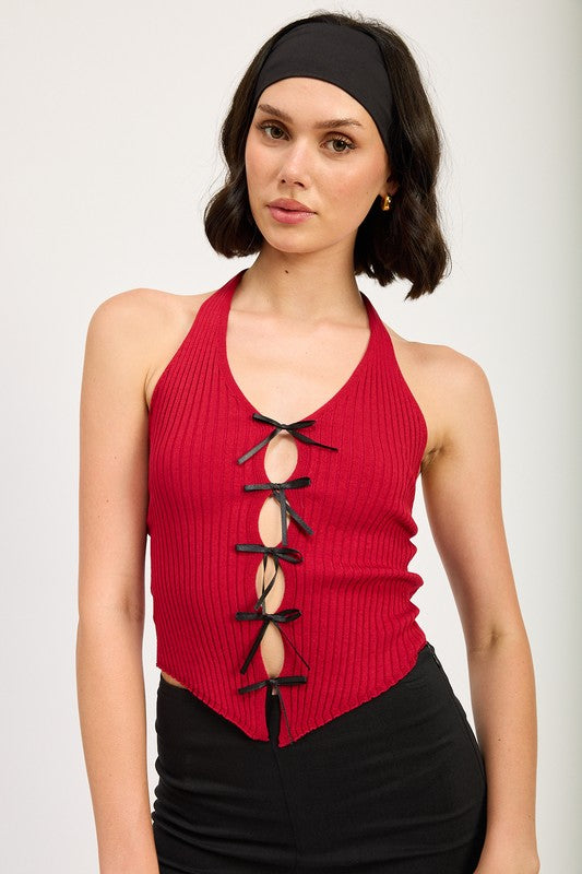 A woman wearing the HALTER NECK TOP WITH BOW DETAILS, a red halter top with black ribbon ties made from 70% viscose and 30% nylon, along with a black headband stands in front of a plain background.