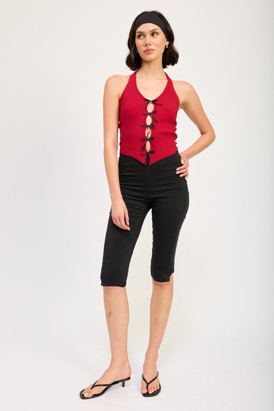 A woman wearing the HALTER NECK TOP WITH BOW DETAILS, a red halter top with black ribbon ties made from 70% viscose and 30% nylon, along with a black headband stands in front of a plain background.