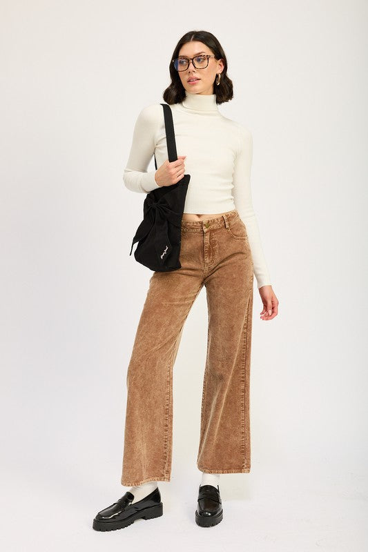 A person wearing a white turtleneck, DISTRASSED CORDUROY PANTS made of 100% cotton in brown, and black loafers with white socks, holding a black shoulder bag, posing in front of a plain background.