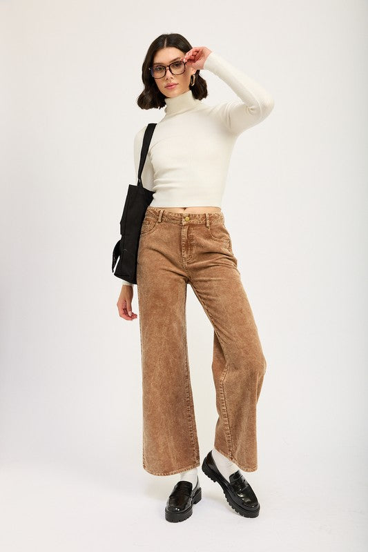 A person wearing a white turtleneck, DISTRASSED CORDUROY PANTS made of 100% cotton in brown, and black loafers with white socks, holding a black shoulder bag, posing in front of a plain background.