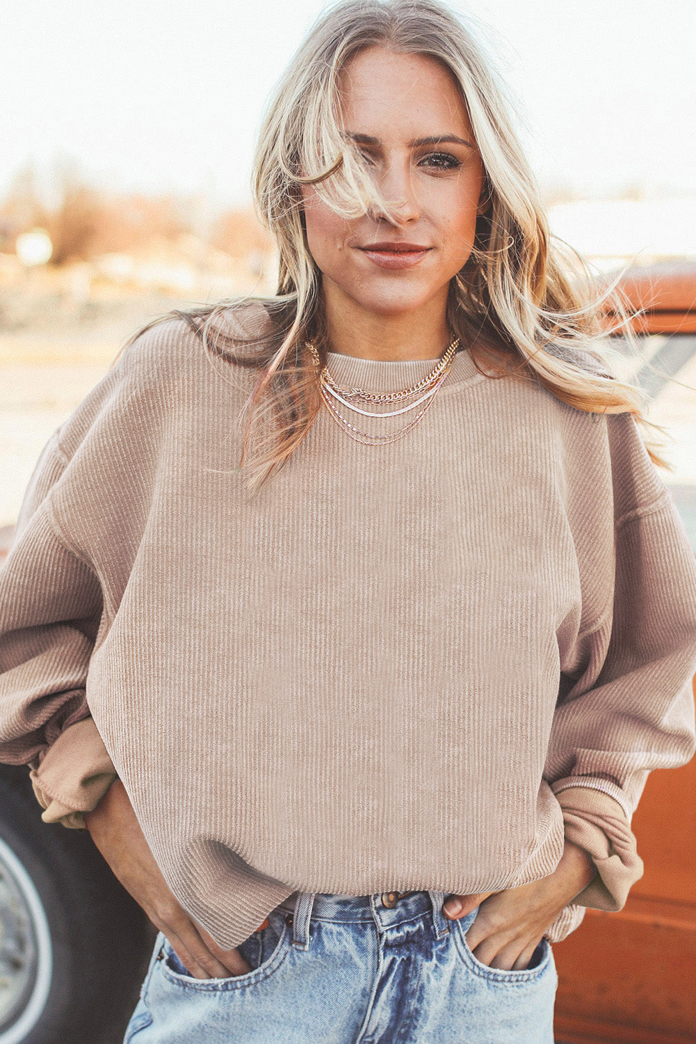 A woman stands outdoors, looking down while touching her hair with one hand. She is wearing an Apricot Ribbed Corduroy Oversized Sweatshirt and white pants, exuding an effortlessly chic and fashionable look.