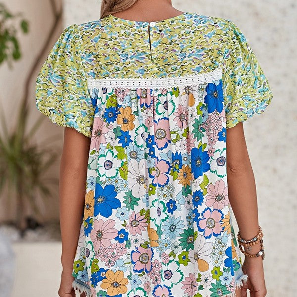 A short-sleeved tunic with bubble sleeves, featuring a lace panel on the chest and a retro pattern of green, blue, pink, and orange flowers on a white background.