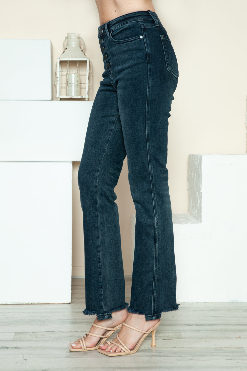 A person is modeling the Judy Blue Full Size Button Fly Hem Destroy Straight Jeans, high-waisted with distressed detailing and frayed hems, paired with light-colored open-toe heels. The background includes white blocks and a light-colored wall.