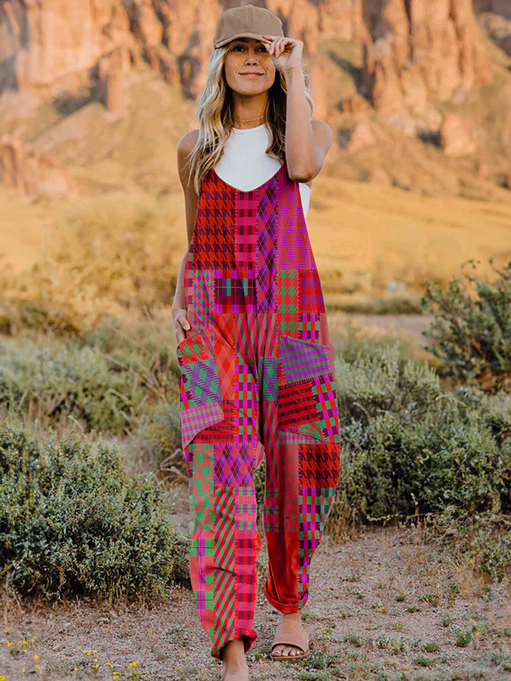 A woman stands outdoors in front of a rocky landscape, wearing the Full Size Printed V-Neck Sleeveless Jumpsuit and a brown brimmed hat. She is smiling and holding the brim of her hat with one hand, exuding the carefree vibe of this vibrant and colorful jumpsuit.