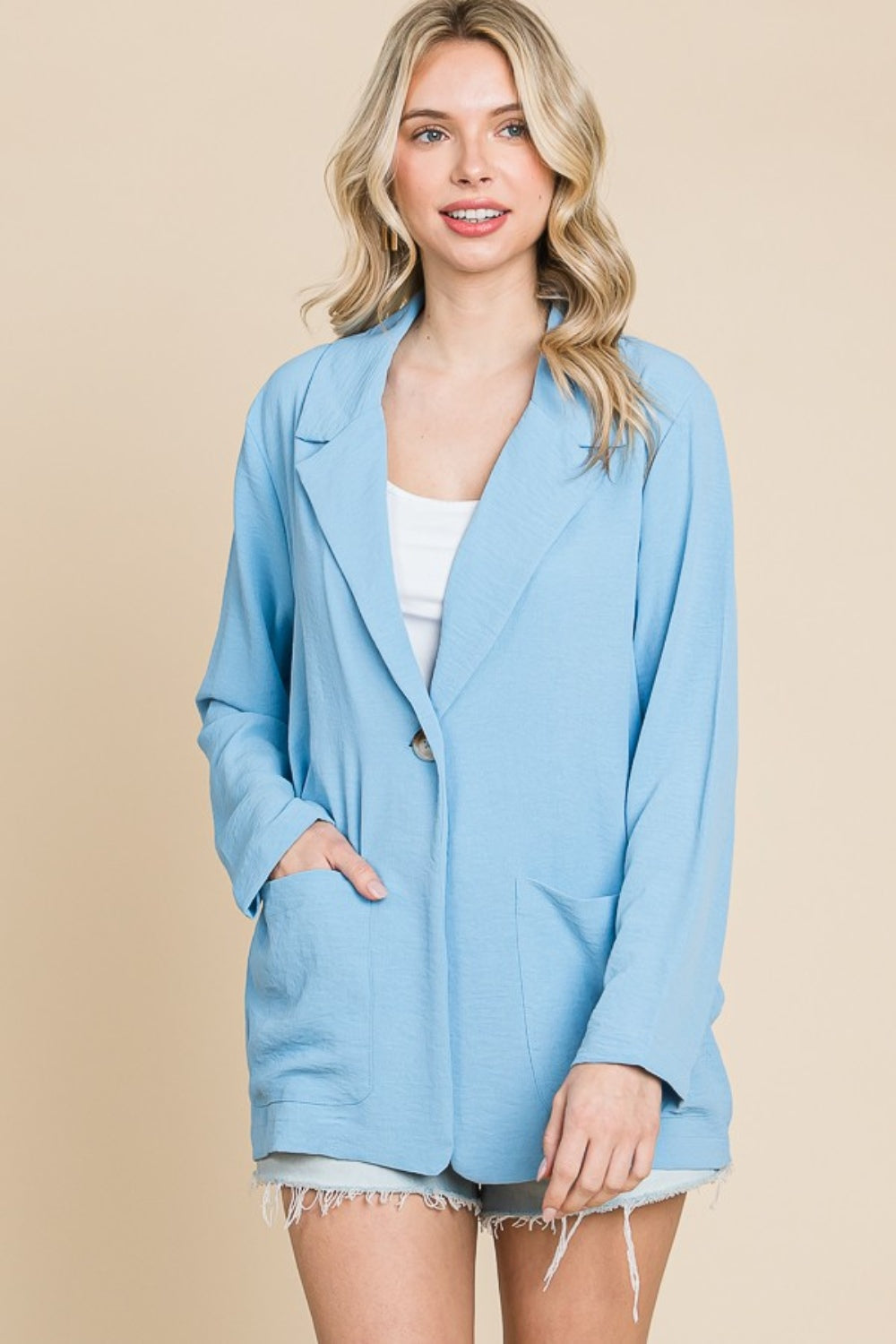 A woman wearing the Culture Code One Button Long Sleeve Blazer with Pockets in light blue over a white top and denim shorts smiles against a beige background, showcasing the versatile charm of her ensemble.