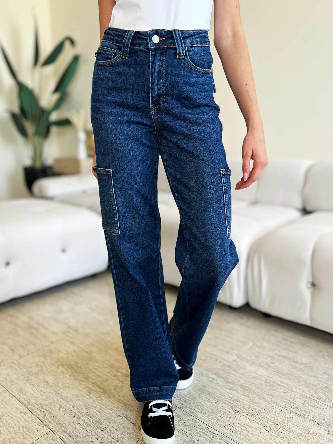 A person wearing Judy Blue Full Size High Waist Straight Cargo Jeans made of durable denim fabric and a white top walks on a wooden floor, with a white sofa and green plant in the background.