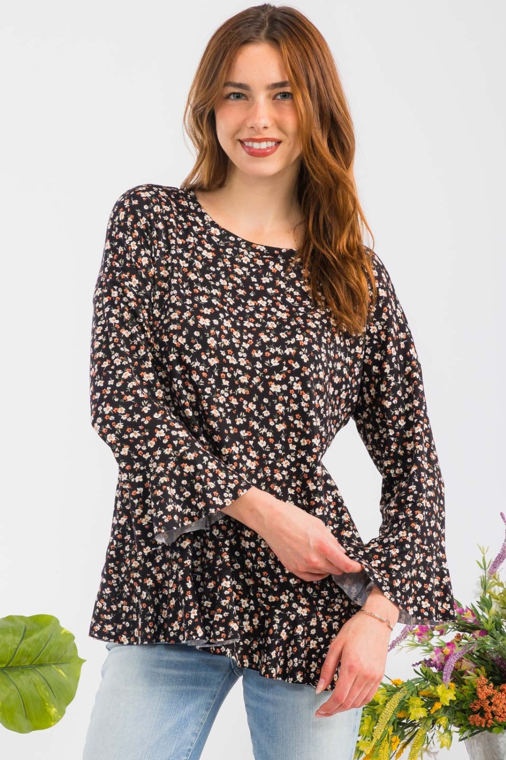 A woman elegantly stands beside colorful flowers against a plain background, embodying feminine fashion in the Celeste Full Size Floral Ruffle Detail Top, which features a round neck and floral patterns, paired with light blue jeans.