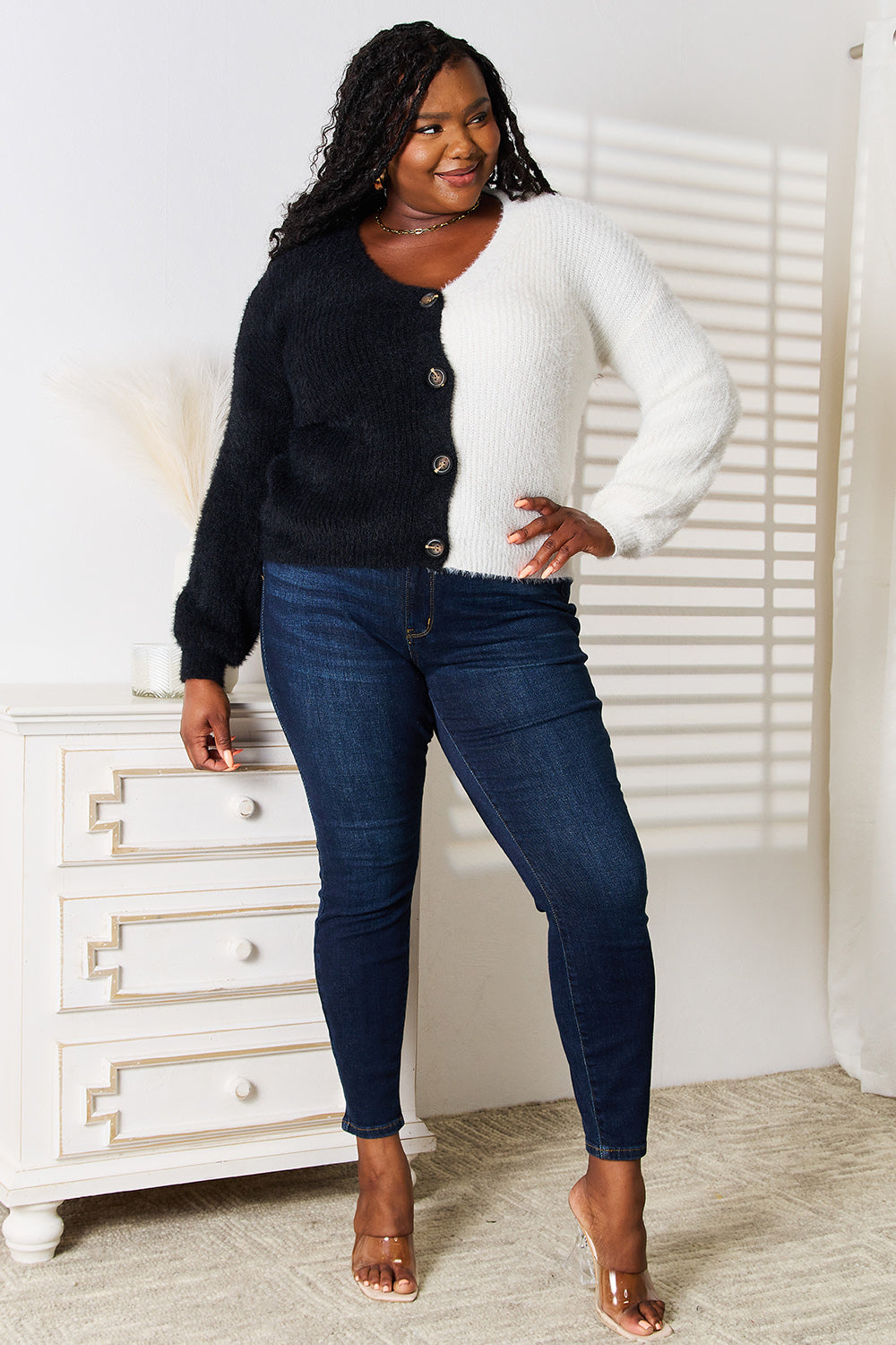 A woman stands with one hand on her hip and the other touching her face. She is wearing the Angel Wings Woven Right Contrast Button-Front V-Neck Cardigan in black and white, paired with jeans. She is smiling.