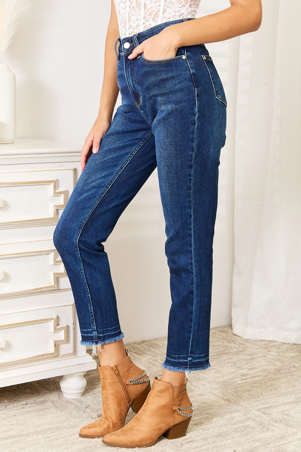 A person wearing Judy Blue Full Size High Waist Released Hem Slit Jeans in indigo wash and a white cropped top stands by a white dresser, showcasing a flattering fit.