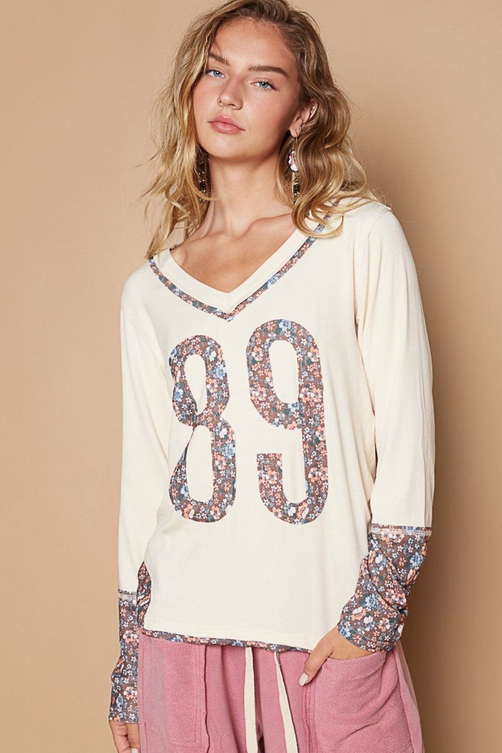 A young woman with blonde hair wears a POL V-Neck Long Sleeve Floral Number Patch T-Shirt, adorned with the number 89 in a floral design on the front and matching floral patterns on the cuffs. She also wears pink pants.