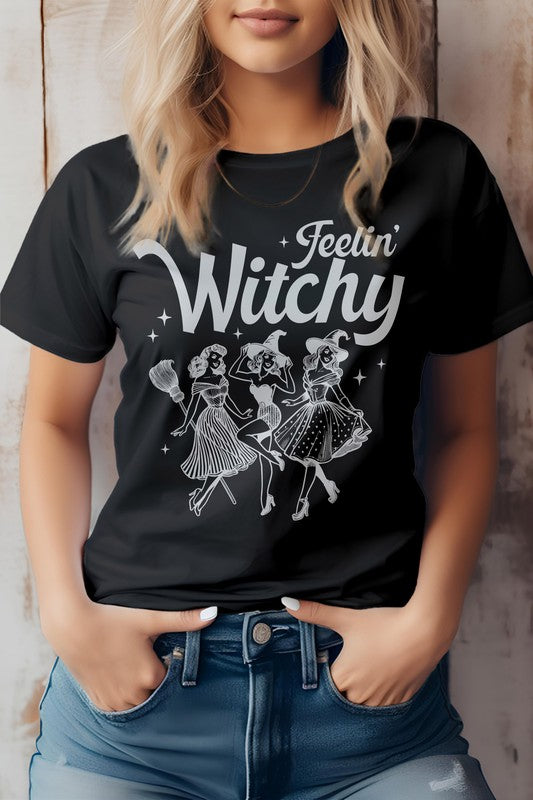 A person is sporting the Feelin' Witchy Halloween Graphic Tee, an unisex pink t-shirt adorned with whimsical illustrations of three dancing witches and the text "Feelin' Witchy." Made from eco-friendly materials, this top effortlessly combines style and sustainability for the spooky season.