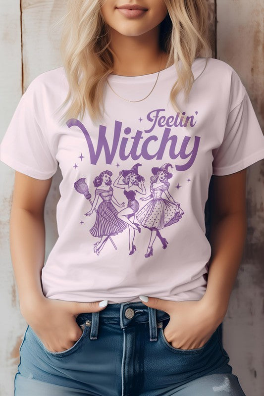 A person is sporting the Feelin' Witchy Halloween Graphic Tee, an unisex pink t-shirt adorned with whimsical illustrations of three dancing witches and the text "Feelin' Witchy." Made from eco-friendly materials, this top effortlessly combines style and sustainability for the spooky season.