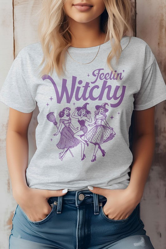 A person is sporting the Feelin' Witchy Halloween Graphic Tee, an unisex pink t-shirt adorned with whimsical illustrations of three dancing witches and the text "Feelin' Witchy." Made from eco-friendly materials, this top effortlessly combines style and sustainability for the spooky season.