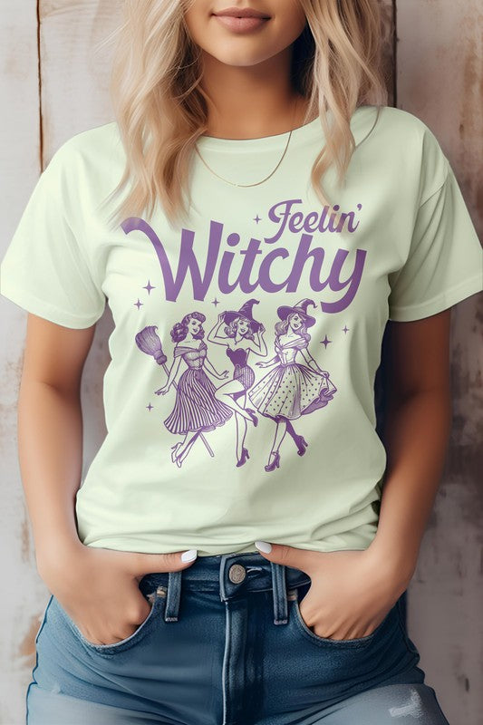 A person is sporting the Feelin' Witchy Halloween Graphic Tee, an unisex pink t-shirt adorned with whimsical illustrations of three dancing witches and the text "Feelin' Witchy." Made from eco-friendly materials, this top effortlessly combines style and sustainability for the spooky season.