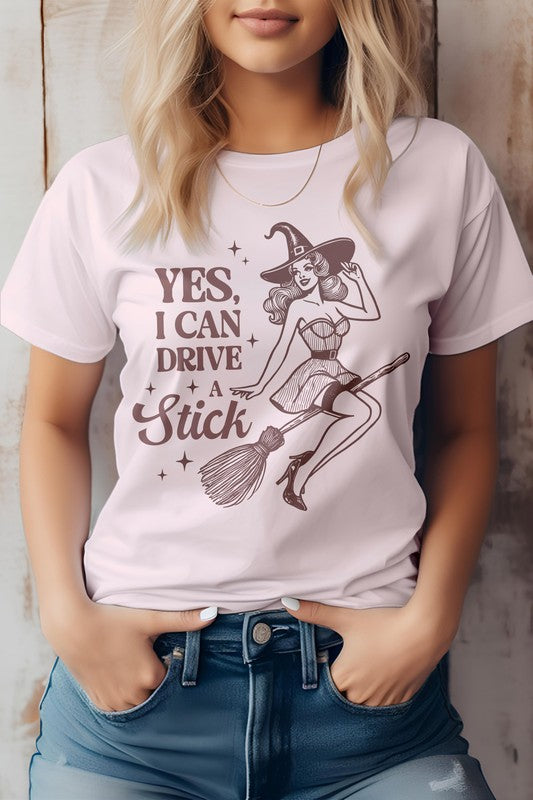 A woman wears the "Yes I Can Drive a Stick, Halloween Graphic Tee," which showcases an illustration of a witch riding a broomstick, complemented by the witty phrase "Yes, I Can Drive a Stick." This Eco-friendly tee combines playful charm with sustainability.