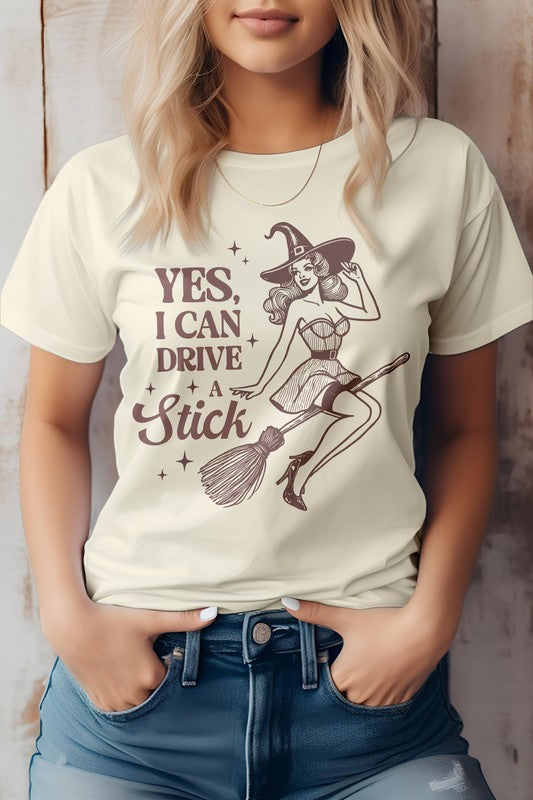 A woman wears the "Yes I Can Drive a Stick, Halloween Graphic Tee," which showcases an illustration of a witch riding a broomstick, complemented by the witty phrase "Yes, I Can Drive a Stick." This Eco-friendly tee combines playful charm with sustainability.