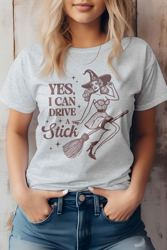 A woman wears the "Yes I Can Drive a Stick, Halloween Graphic Tee," which showcases an illustration of a witch riding a broomstick, complemented by the witty phrase "Yes, I Can Drive a Stick." This Eco-friendly tee combines playful charm with sustainability.