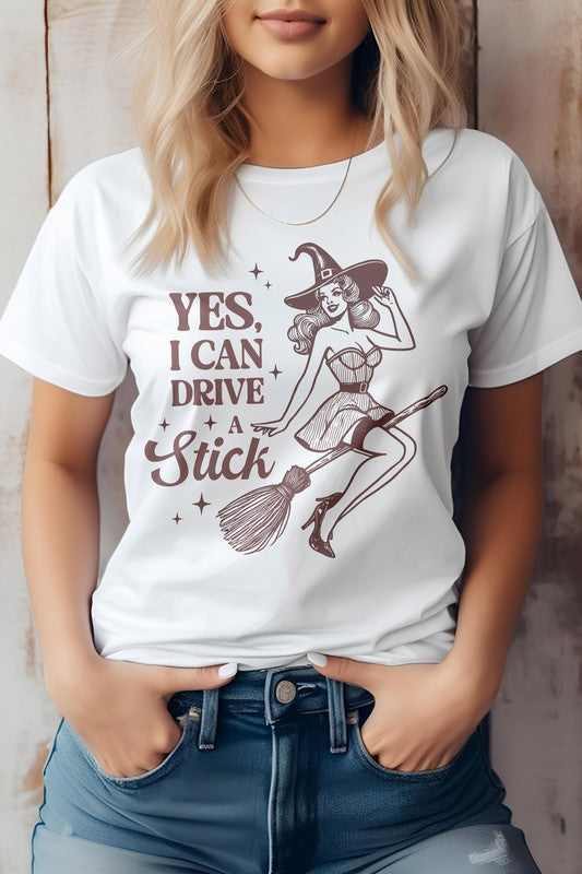 A woman wears the "Yes I Can Drive a Stick, Halloween Graphic Tee," which showcases an illustration of a witch riding a broomstick, complemented by the witty phrase "Yes, I Can Drive a Stick." This Eco-friendly tee combines playful charm with sustainability.