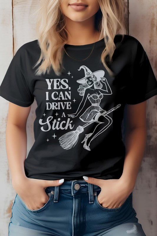 A woman wears the "Yes I Can Drive a Stick, Halloween Graphic Tee," which showcases an illustration of a witch riding a broomstick, complemented by the witty phrase "Yes, I Can Drive a Stick." This Eco-friendly tee combines playful charm with sustainability.