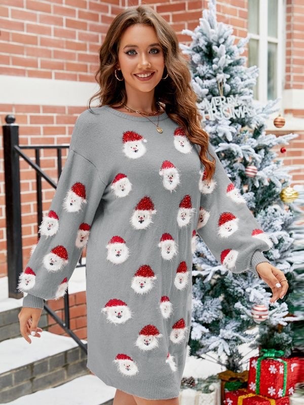 A woman smiles while wearing the Santa sweater dress, featuring pink fabric adorned with Santa Claus faces. She stands in front of a decorated Christmas tree and a brick building, showcasing her holiday fashion.