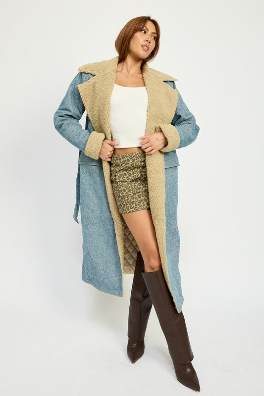 A woman stands wearing the IMA9409J-1 Denim Maxi Coat with tan accents made of imported polyester, brown knee-high boots, a white cotton top, and sunglasses resting on her head.