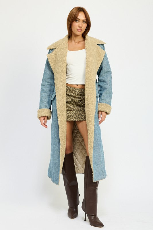 A woman stands wearing the IMA9409J-1 Denim Maxi Coat with tan accents made of imported polyester, brown knee-high boots, a white cotton top, and sunglasses resting on her head.