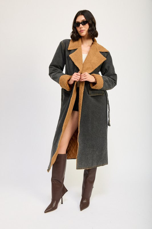 A woman stands wearing the IMA9409J-1 Denim Maxi Coat with tan accents made of imported polyester, brown knee-high boots, a white cotton top, and sunglasses resting on her head.