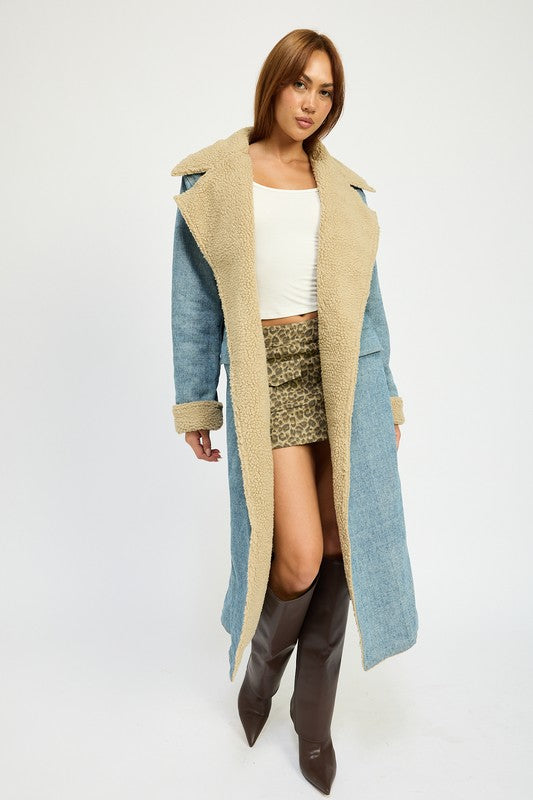 A woman stands wearing the IMA9409J-1 Denim Maxi Coat with tan accents made of imported polyester, brown knee-high boots, a white cotton top, and sunglasses resting on her head.