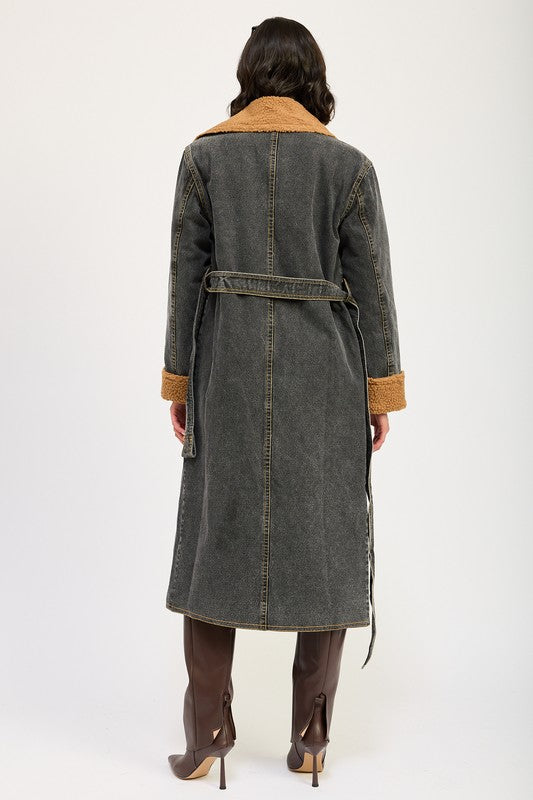 A woman stands wearing the IMA9409J-1 Denim Maxi Coat with tan accents made of imported polyester, brown knee-high boots, a white cotton top, and sunglasses resting on her head.