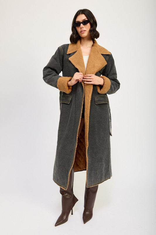 A woman stands wearing the IMA9409J-1 Denim Maxi Coat with tan accents made of imported polyester, brown knee-high boots, a white cotton top, and sunglasses resting on her head.