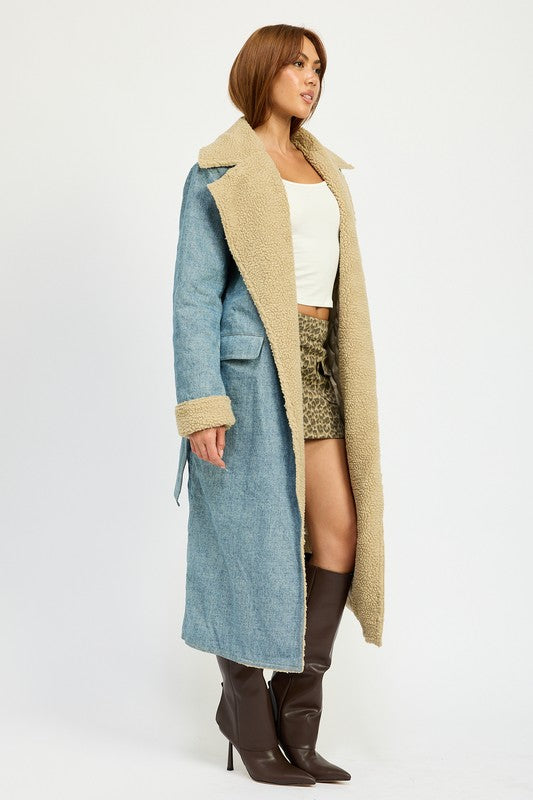 A woman stands wearing the IMA9409J-1 Denim Maxi Coat with tan accents made of imported polyester, brown knee-high boots, a white cotton top, and sunglasses resting on her head.