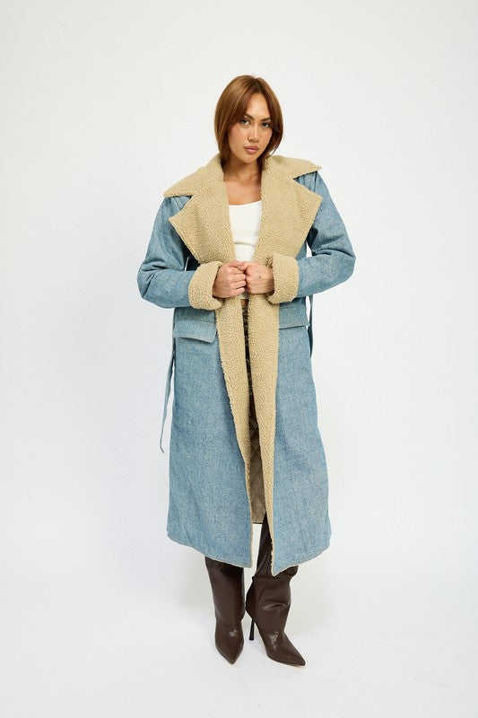 A woman stands wearing the IMA9409J-1 Denim Maxi Coat with tan accents made of imported polyester, brown knee-high boots, a white cotton top, and sunglasses resting on her head.