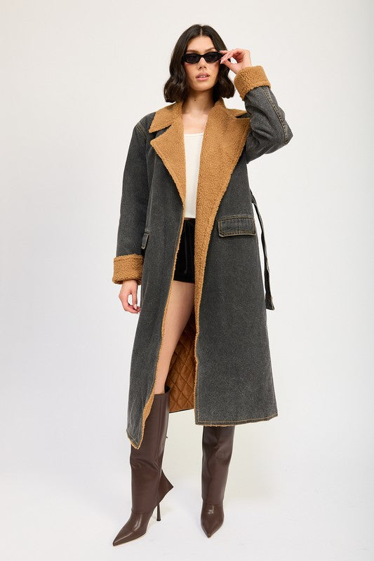 A woman stands wearing the IMA9409J-1 Denim Maxi Coat with tan accents made of imported polyester, brown knee-high boots, a white cotton top, and sunglasses resting on her head.