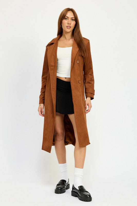 A person wearing a long brown SUEDE TRENCH COAT over a white top and black skirt, both featuring spandex for a perfect size & fit, stands against a plain background. They are also wearing black shoes and white socks.