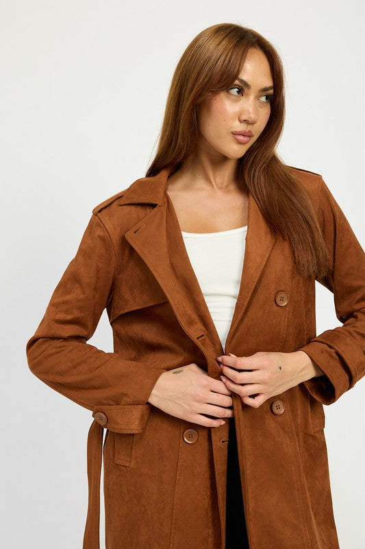 A person wearing a long brown SUEDE TRENCH COAT over a white top and black skirt, both featuring spandex for a perfect size & fit, stands against a plain background. They are also wearing black shoes and white socks.