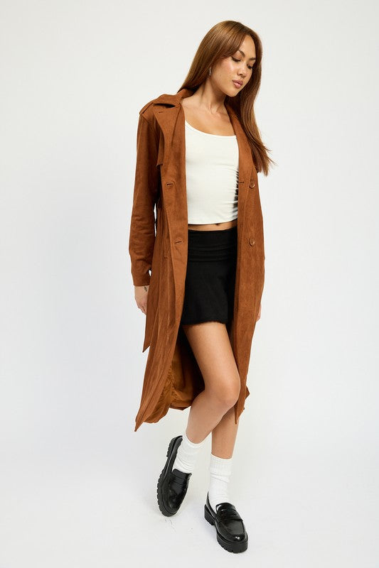 A person wearing a long brown SUEDE TRENCH COAT over a white top and black skirt, both featuring spandex for a perfect size & fit, stands against a plain background. They are also wearing black shoes and white socks.