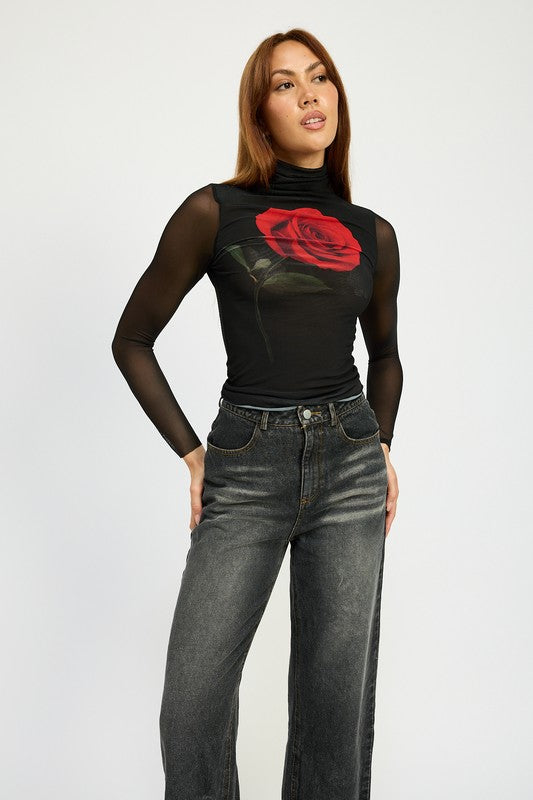 A person with long hair wearing a black mesh top adorned with a rose design, high-waisted WASHED DENIM PANTS WITH FOLD OVER DETAIL in dark color, and red pointed shoes stands against a plain background. The outfit includes imported, size small pieces made from 100% cotton.