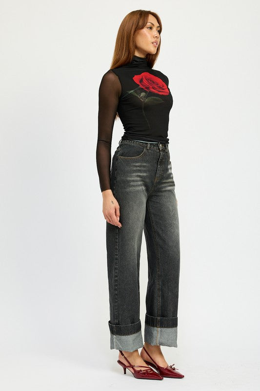 A person with long hair wearing a black mesh top adorned with a rose design, high-waisted WASHED DENIM PANTS WITH FOLD OVER DETAIL in dark color, and red pointed shoes stands against a plain background. The outfit includes imported, size small pieces made from 100% cotton.