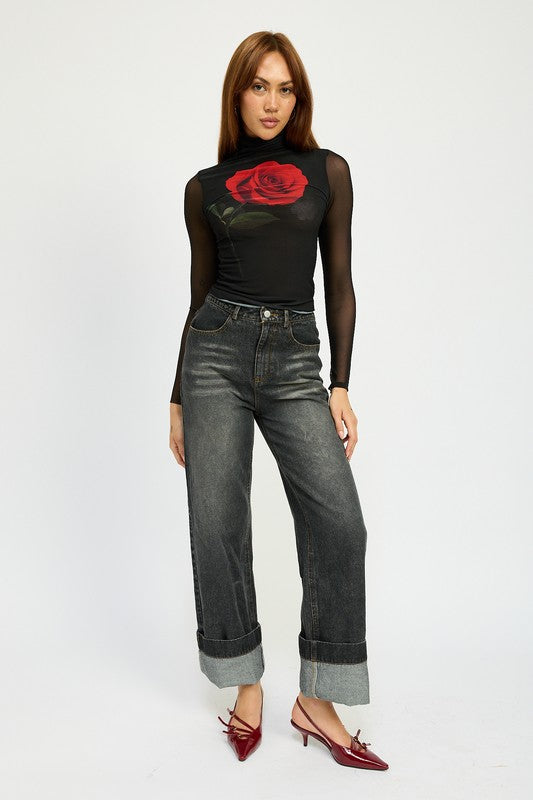 A person with long hair wearing a black mesh top adorned with a rose design, high-waisted WASHED DENIM PANTS WITH FOLD OVER DETAIL in dark color, and red pointed shoes stands against a plain background. The outfit includes imported, size small pieces made from 100% cotton.