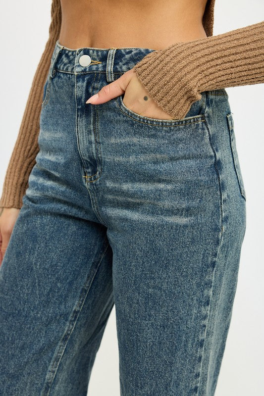 A person with long hair wearing a black mesh top adorned with a rose design, high-waisted WASHED DENIM PANTS WITH FOLD OVER DETAIL in dark color, and red pointed shoes stands against a plain background. The outfit includes imported, size small pieces made from 100% cotton.