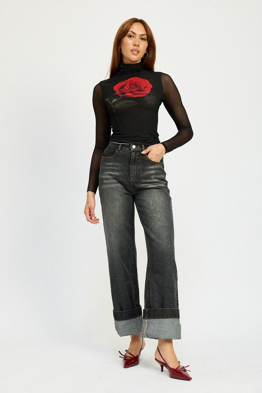 A person with long hair wearing a black mesh top adorned with a rose design, high-waisted WASHED DENIM PANTS WITH FOLD OVER DETAIL in dark color, and red pointed shoes stands against a plain background. The outfit includes imported, size small pieces made from 100% cotton.