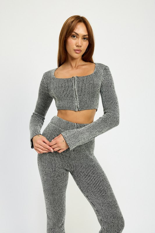 A person with long hair is wearing a grey, ribbed SQUARE NECK CROPPED TOP WITH ZIPPER, made from 60% cotton and paired with high-waisted pants. They are standing and looking directly at the camera. The model wears size small.