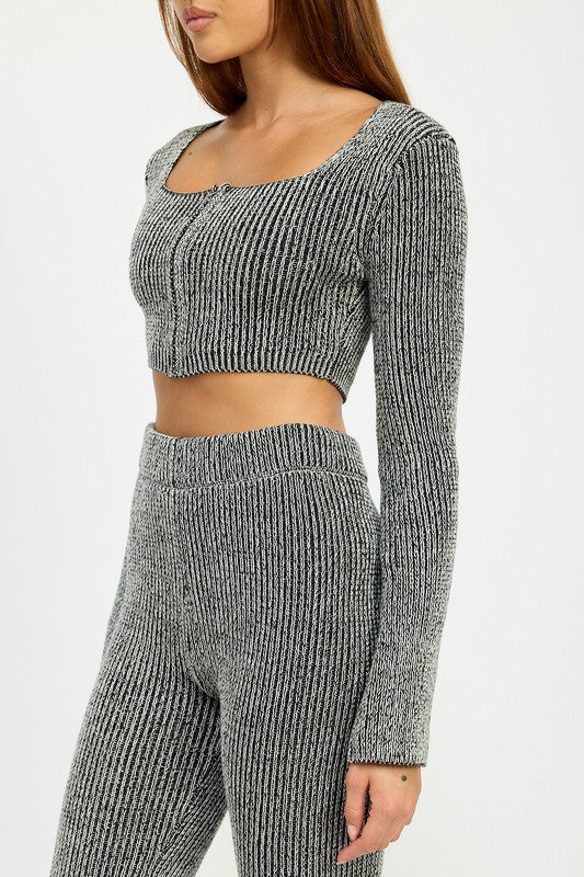 A person with long hair is wearing a grey, ribbed SQUARE NECK CROPPED TOP WITH ZIPPER, made from 60% cotton and paired with high-waisted pants. They are standing and looking directly at the camera. The model wears size small.