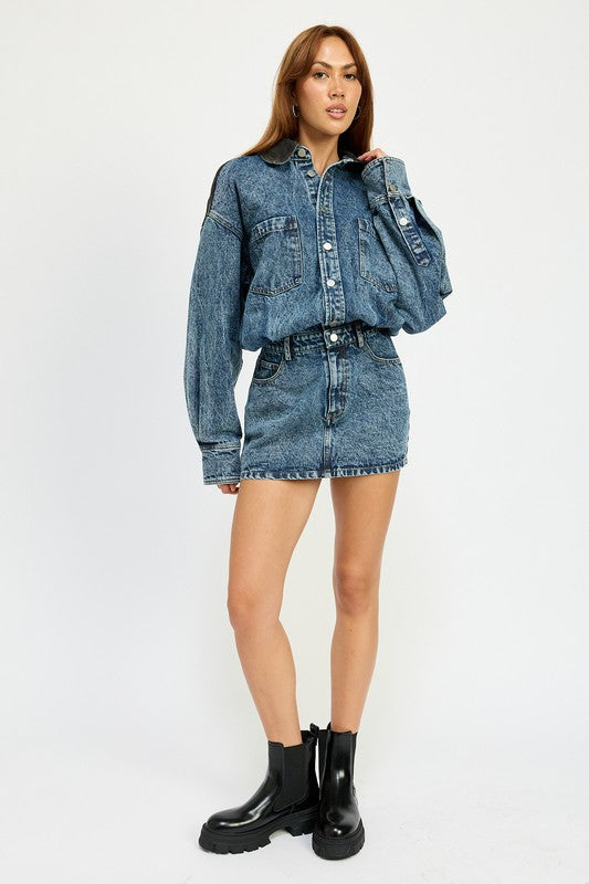 A person stands in a narrow doorway wearing an imported Button Up Denim Mini Dress and white knee-high boots, with one arm resting on the door frame. The 100% cotton ensemble exudes effortless style and comfort.