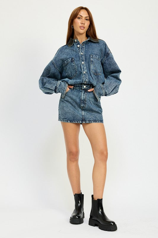 A person stands in a narrow doorway wearing an imported Button Up Denim Mini Dress and white knee-high boots, with one arm resting on the door frame. The 100% cotton ensemble exudes effortless style and comfort.