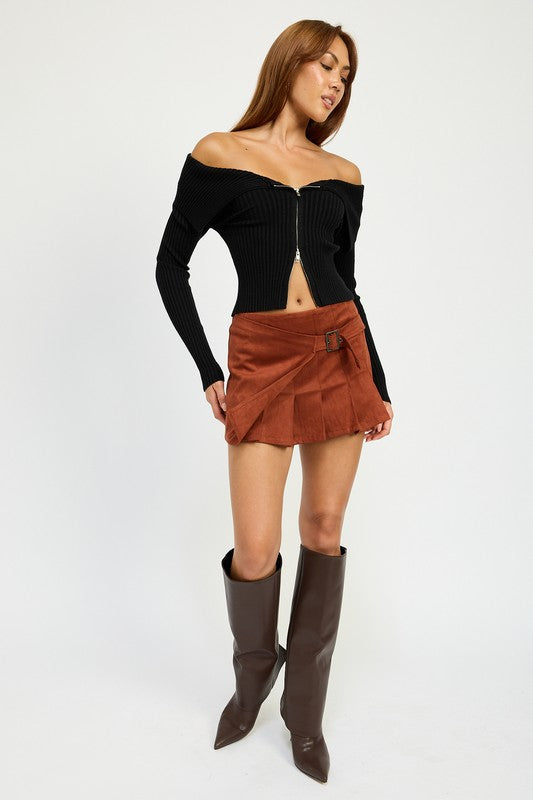 A woman stands against a white background, wearing an off-the-shoulder black top made of polyester and spandex, the WRAPPED BUCKLE BELT MINI SKIRT in orange, and brown knee-high boots. She is dressed in size small.