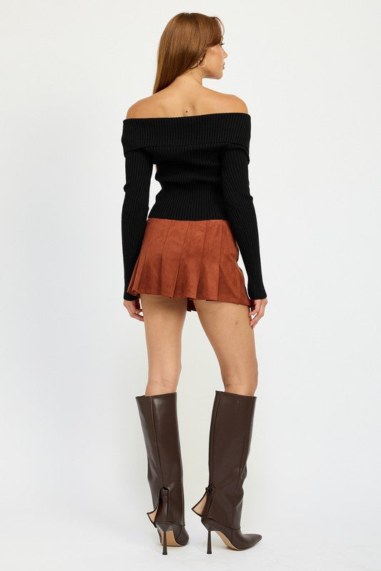 A woman stands against a white background, wearing an off-the-shoulder black top made of polyester and spandex, the WRAPPED BUCKLE BELT MINI SKIRT in orange, and brown knee-high boots. She is dressed in size small.